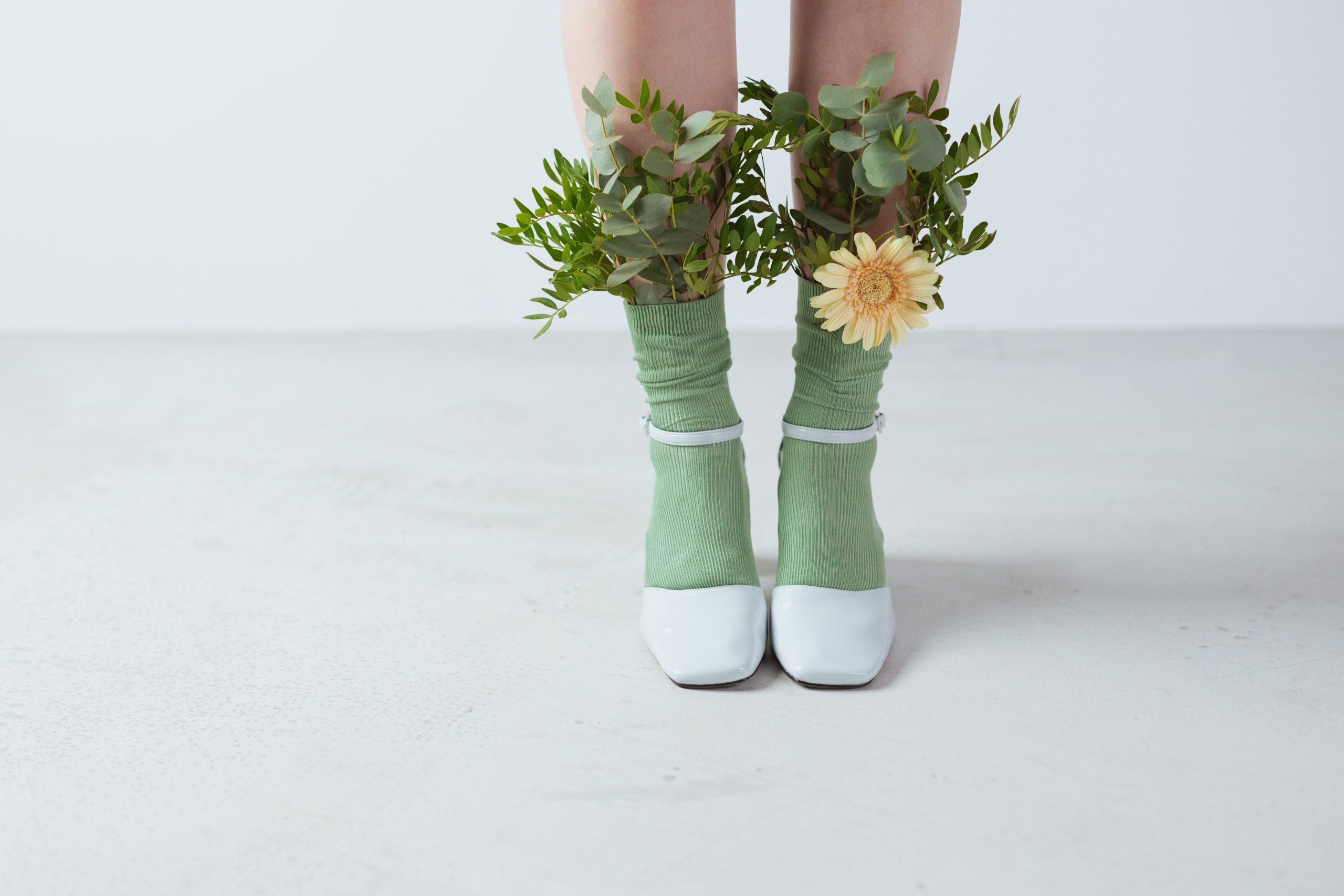 Artsy photo of woman in green socks and white heels with bunches of flowers sticking out the top 